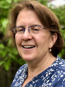 Ellen J. DeBenedetti, woman with fair skin, light brown hair, glasses and a blue shirt with white flowers